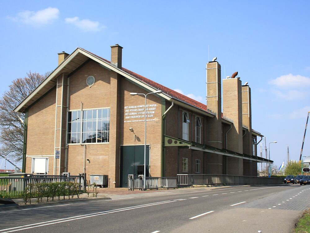 Bakstenen gebouw van Gemaal Vissering op Urk onder een blauwe lucht, met op de achtergrond masten van schepen in de haven